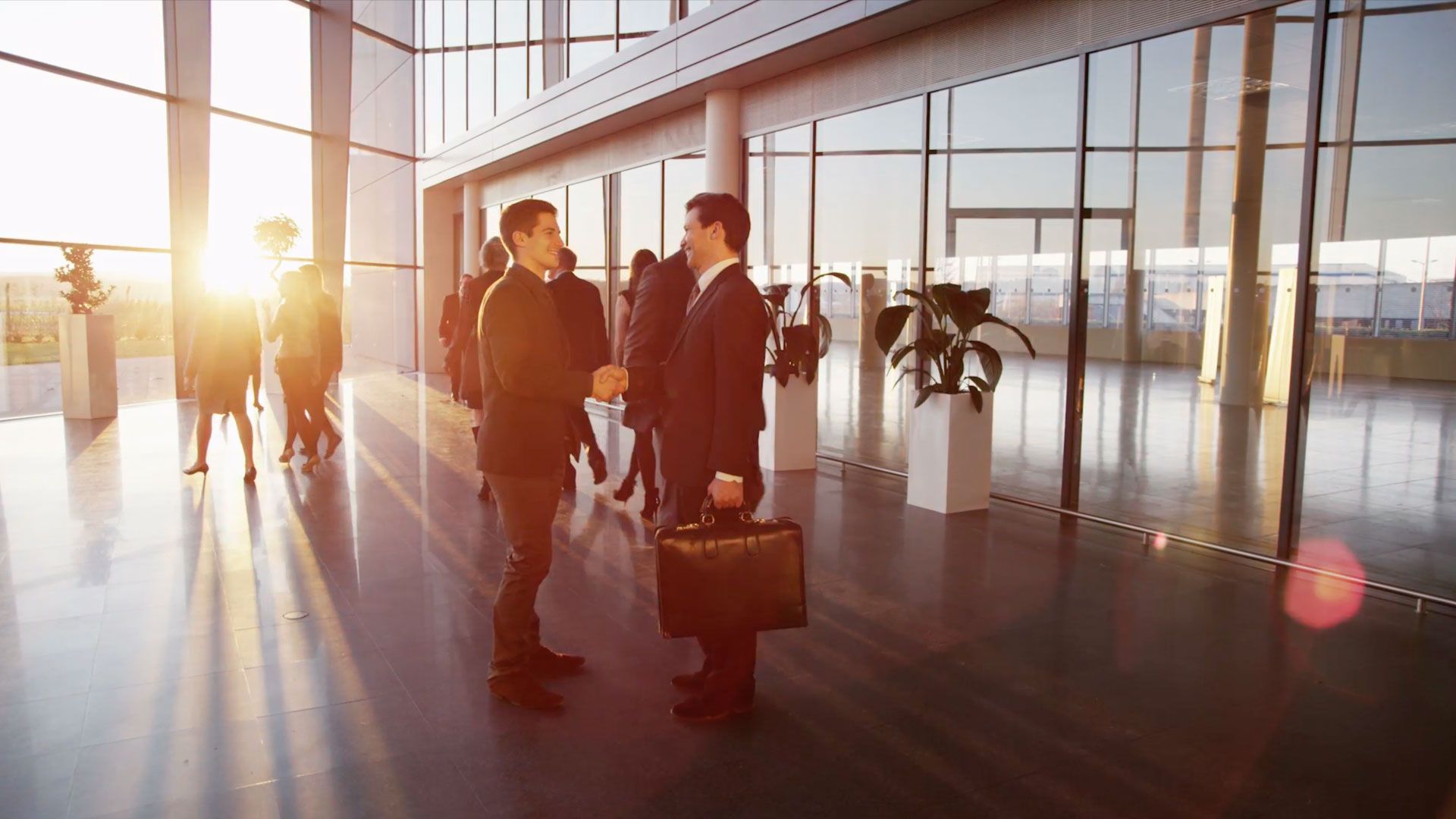 Two businessmen shaking hands in a modern office lobby at sunset.
